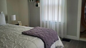 A bedroom with a large bed, a bedside table with a lamp, a window with sheer curtains, and a small plant on the wall. - Cozy Blue Cottage With Access To All Sports Lake.  (Greenville)