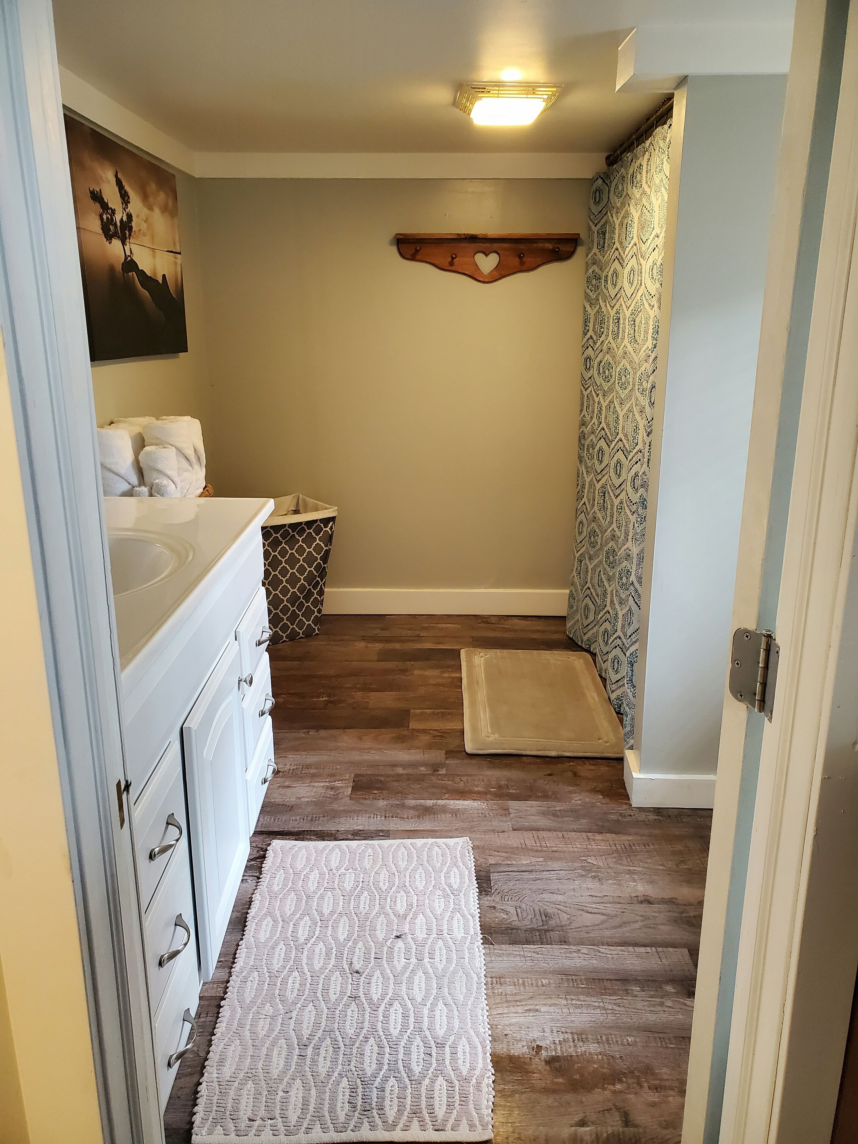 Un baño con piso de madera, lavamanos blanco y un perchero para toallas. Hay una alfombra estampada en el piso y una cortina de ducha con diseño geométrico.