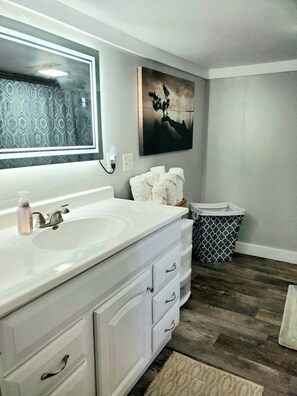 A modern bathroom with a large mirror, white vanity, and a painting on the wall. - Cozy Blue Cottage With Access To All Sports Lake.  (Greenville)