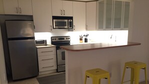 Una cocina moderna con un refrigerador de acero inoxidable, un microondas, un horno y un tostador. Hay dos taburetes amarillos y una pequeña alfombra en el suelo.