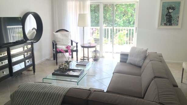 Una sala de estar con un sofá, una mesa de centro, una silla, un mueble para televisión y una ventana con vista a follaje.