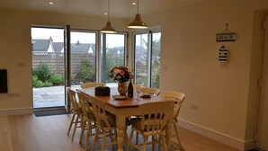 A dining area with a wooden table and wicker chairs, a vase with flowers, and a view of the garden through glass doors.