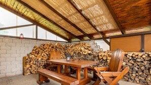 Un área de estar al aire libre de madera rústica, con una mesa, bancos, una pila de leña y una pared de ladrillo.