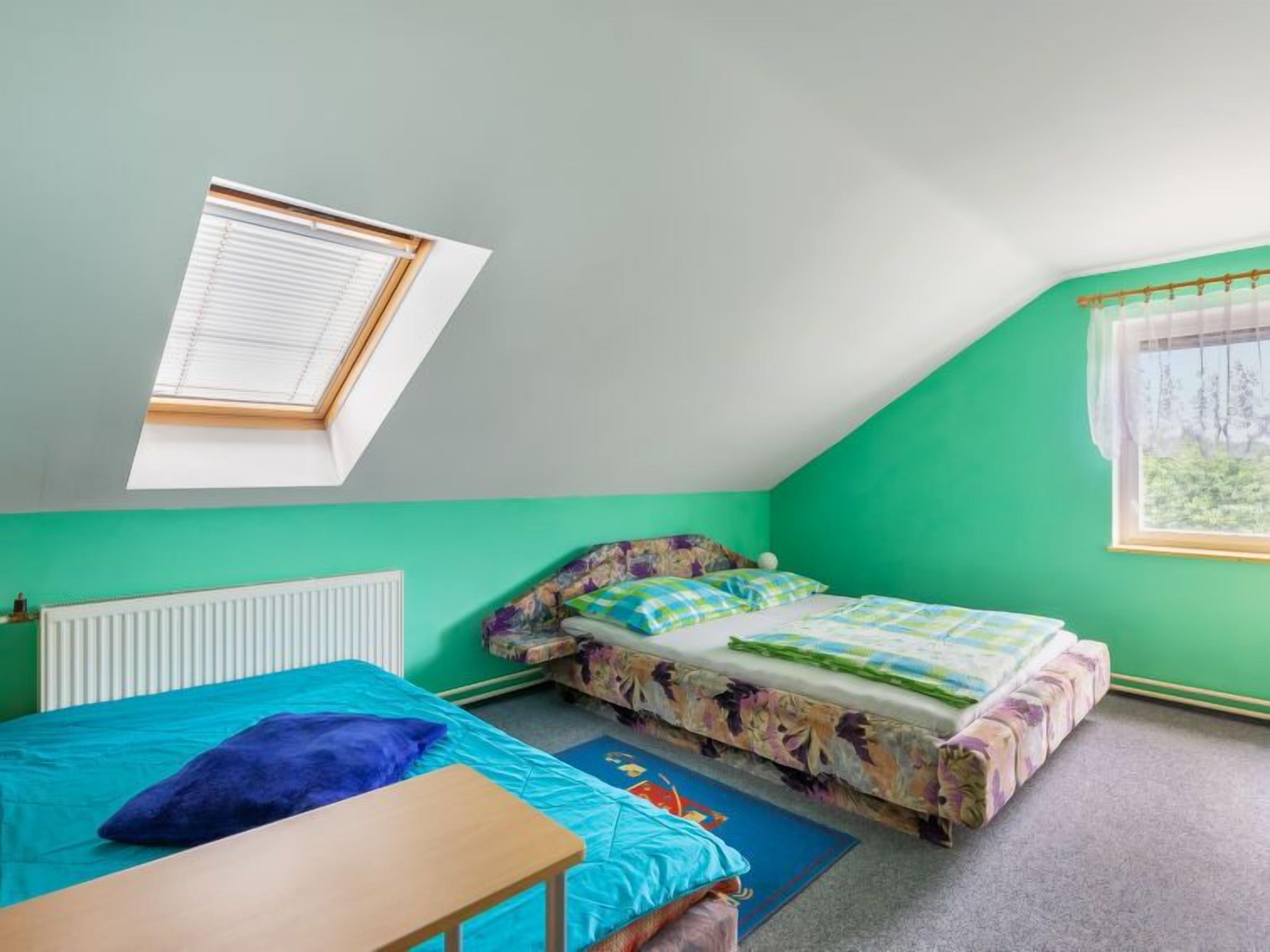 Une chambre avec un plafond en pente, un lit recouvert d’une literie à motifs floraux, une petite table et une fenêtre donnant sur un paysage verdoyant.