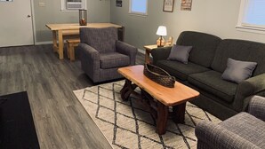 A cozy living room with a gray couch, a wooden coffee table, and a plaid armchair. There is a dining table with chairs, a fireplace, and a clock on the wall. - Hilltop Cabin at Wood Creek Lake (London)