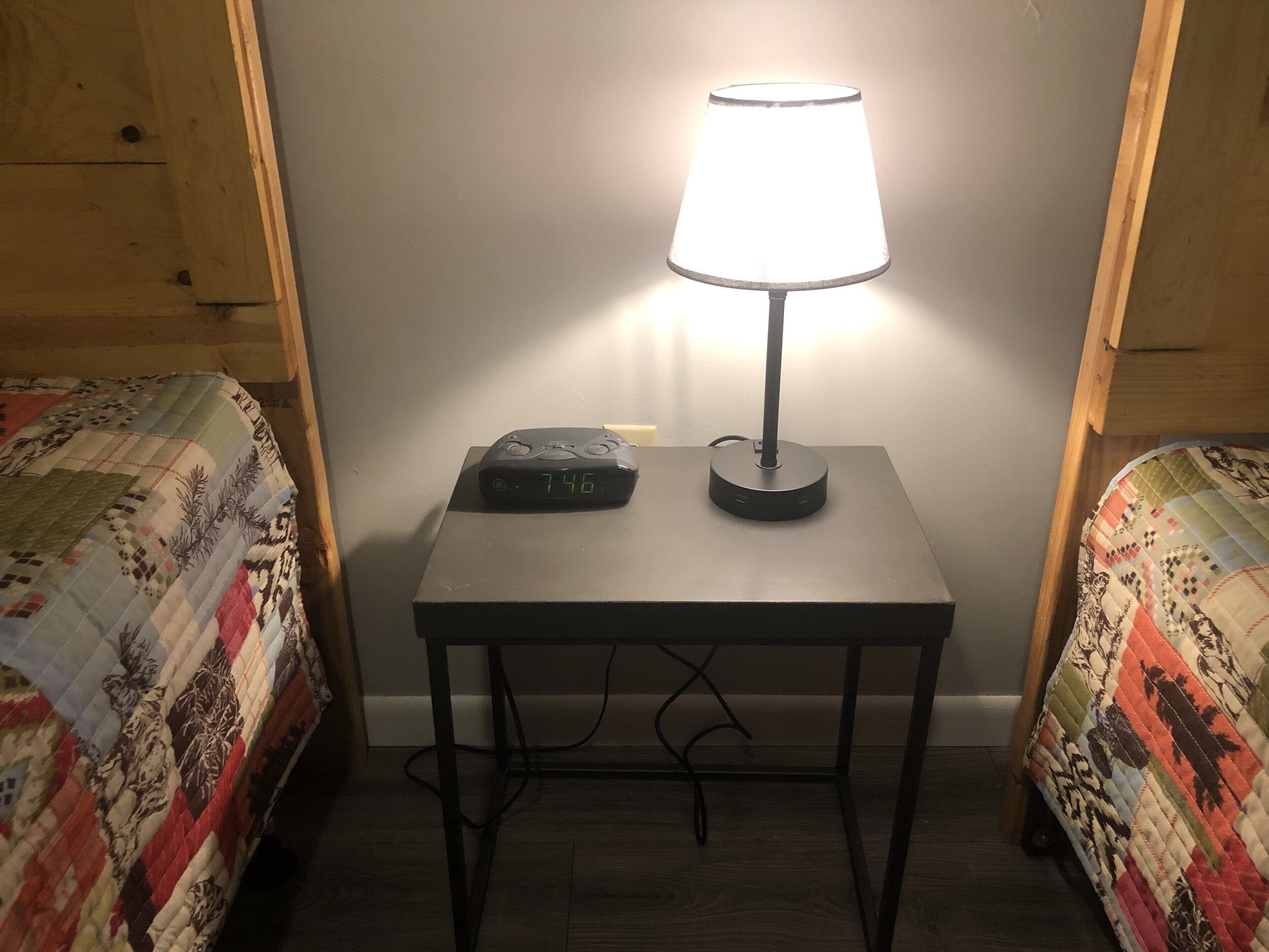 A small table with a lamp and a clock in a room with two beds.