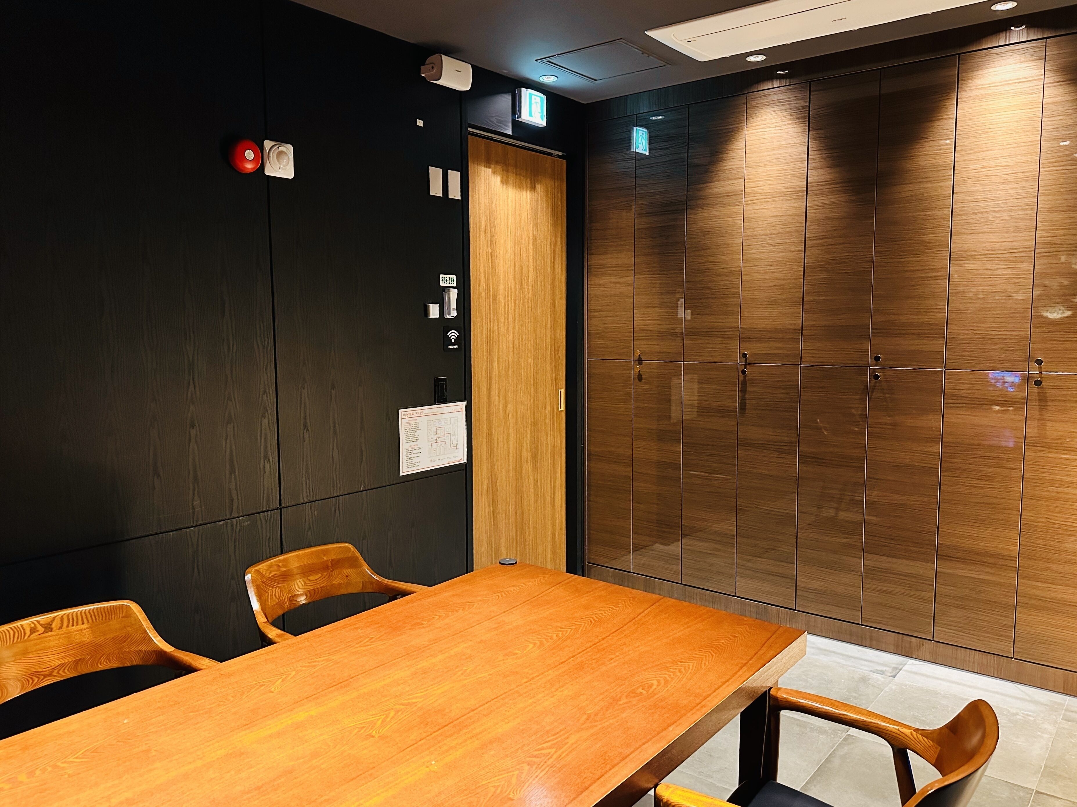 Un salón de conferencias con una mesa y sillas de madera, una pared negra con detector de incendios y un armario de madera con espejo.