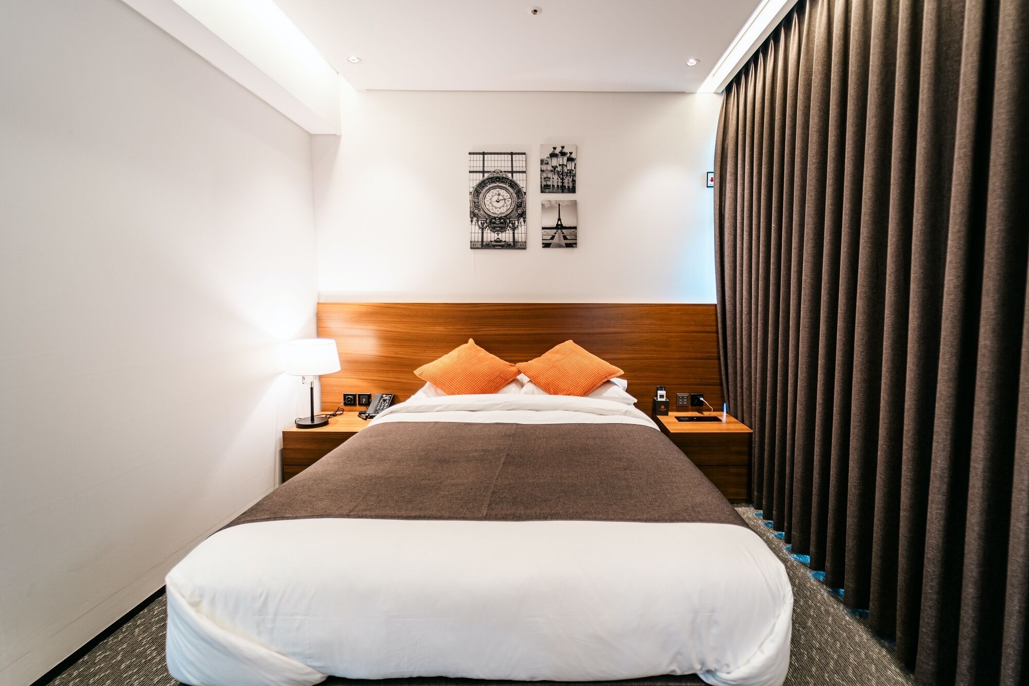 Camera d'hotel con un letto grande, una testiera in legno, due comodini con lampade e un tavolino da notte, e vista sulla Torre Eiffel dalla finestra.
