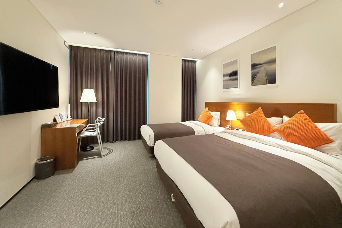Ett hotellrum med två sängar, ett skrivbord, en stol, en TV och ett fönster med gardiner.