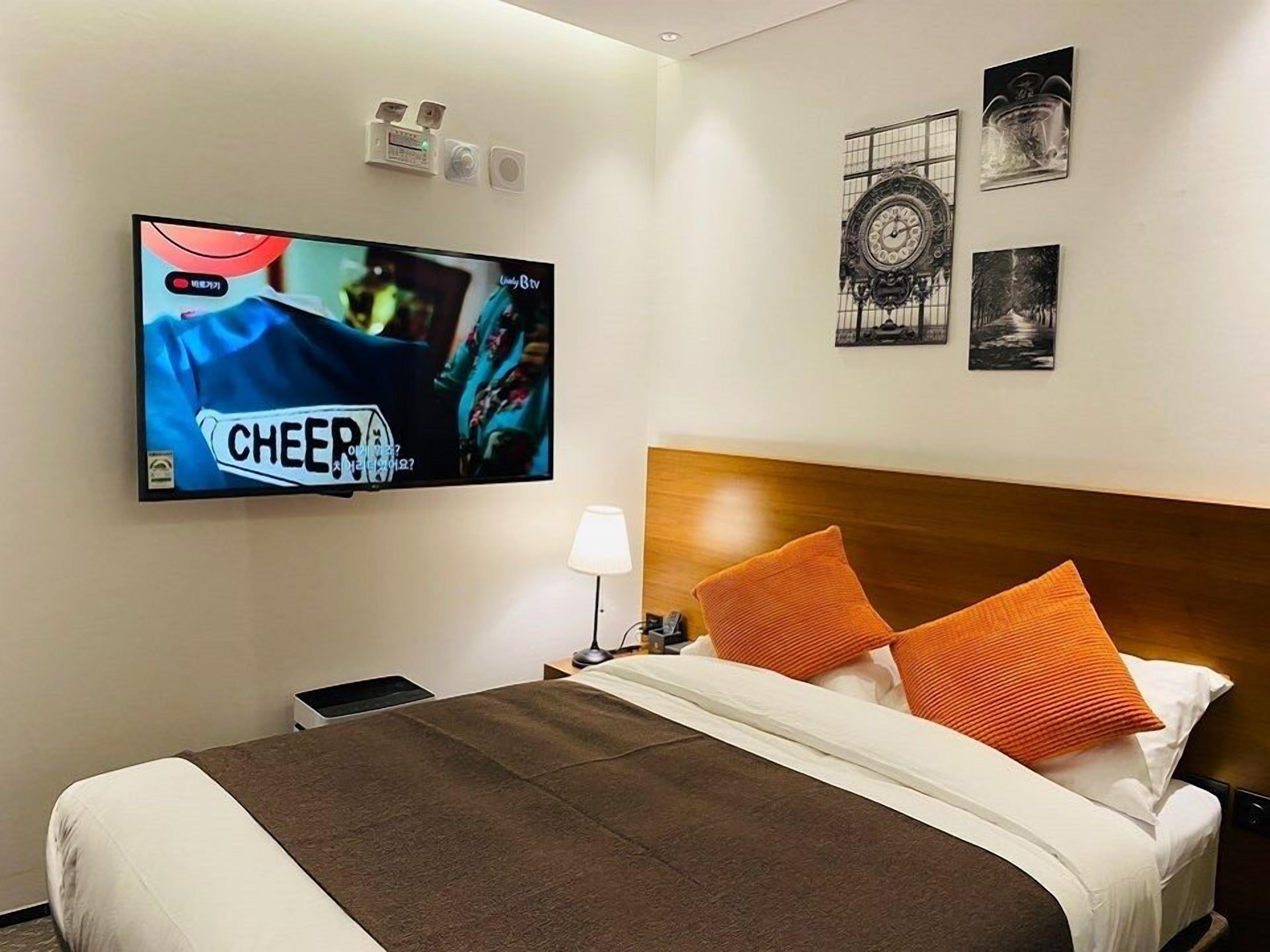 Une chambre d’hôtel dotée d’un grand téléviseur à écran plat fixé au mur, d’une tête de lit en bois, de deux oreillers orange, d’un plaid marron et d’une table de chevet avec une lampe.
