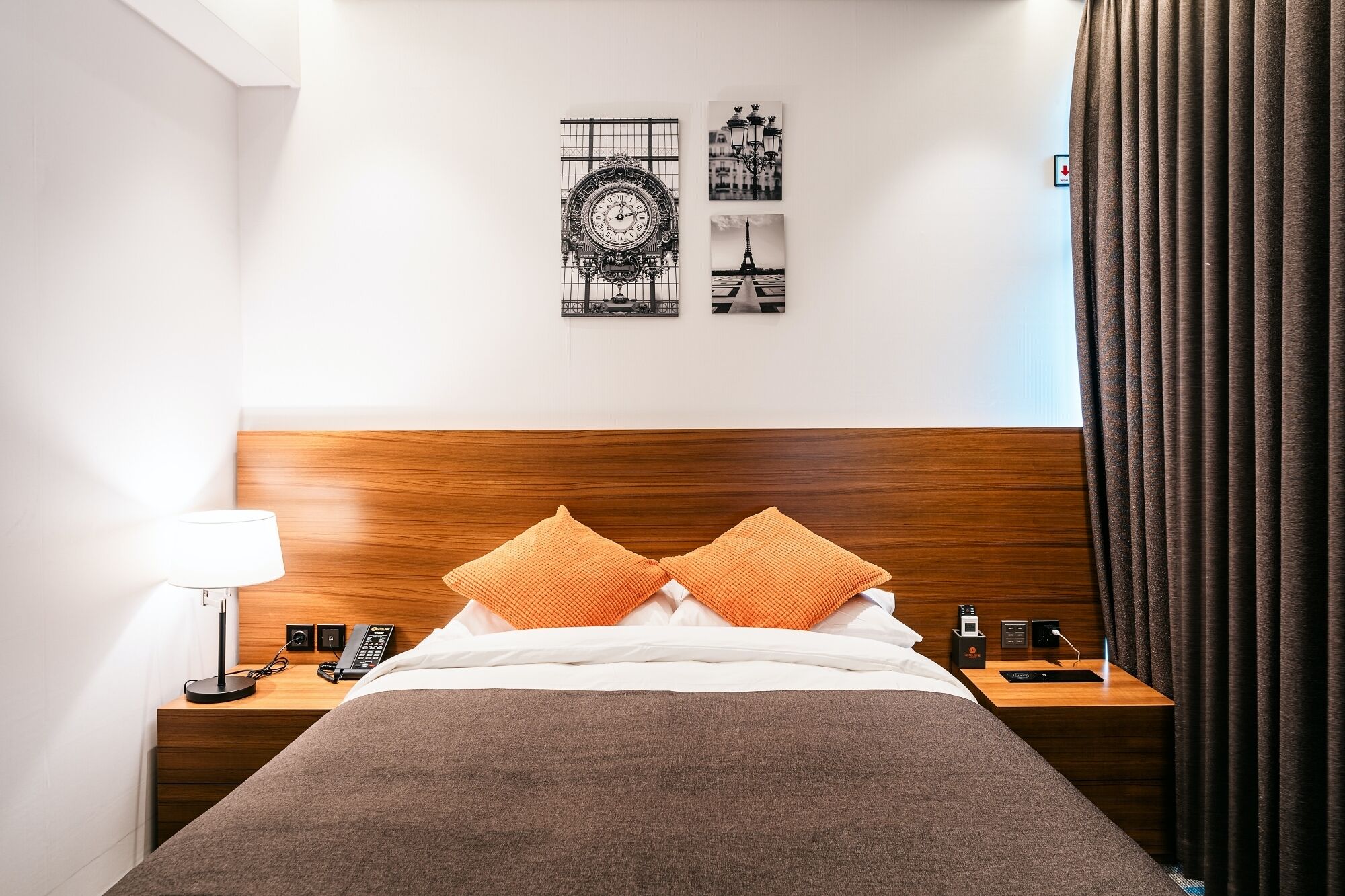 A hotel room with a wooden headboard, two orange pillows, a white bedspread, a nightstand with a lamp, a phone, and a remote control.
