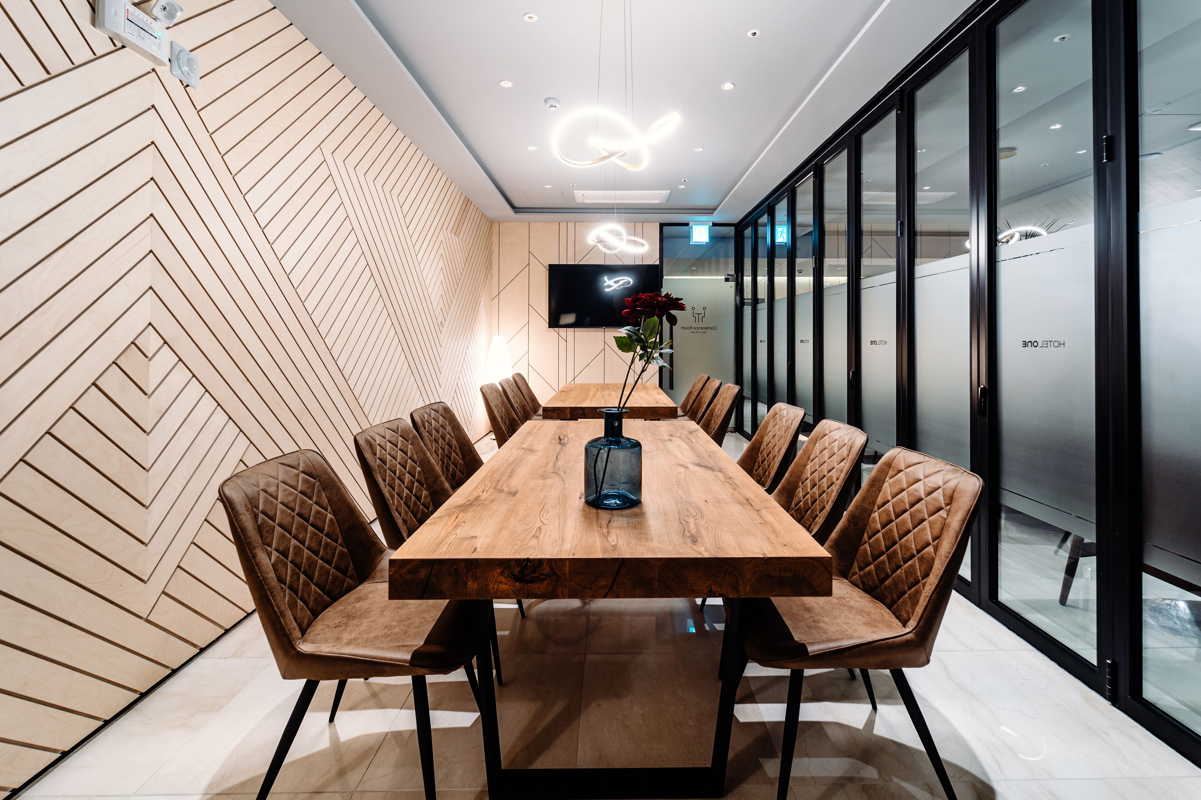 A conference room with a long wooden table, cushioned chairs, a flat-screen TV, and a modern light fixture.