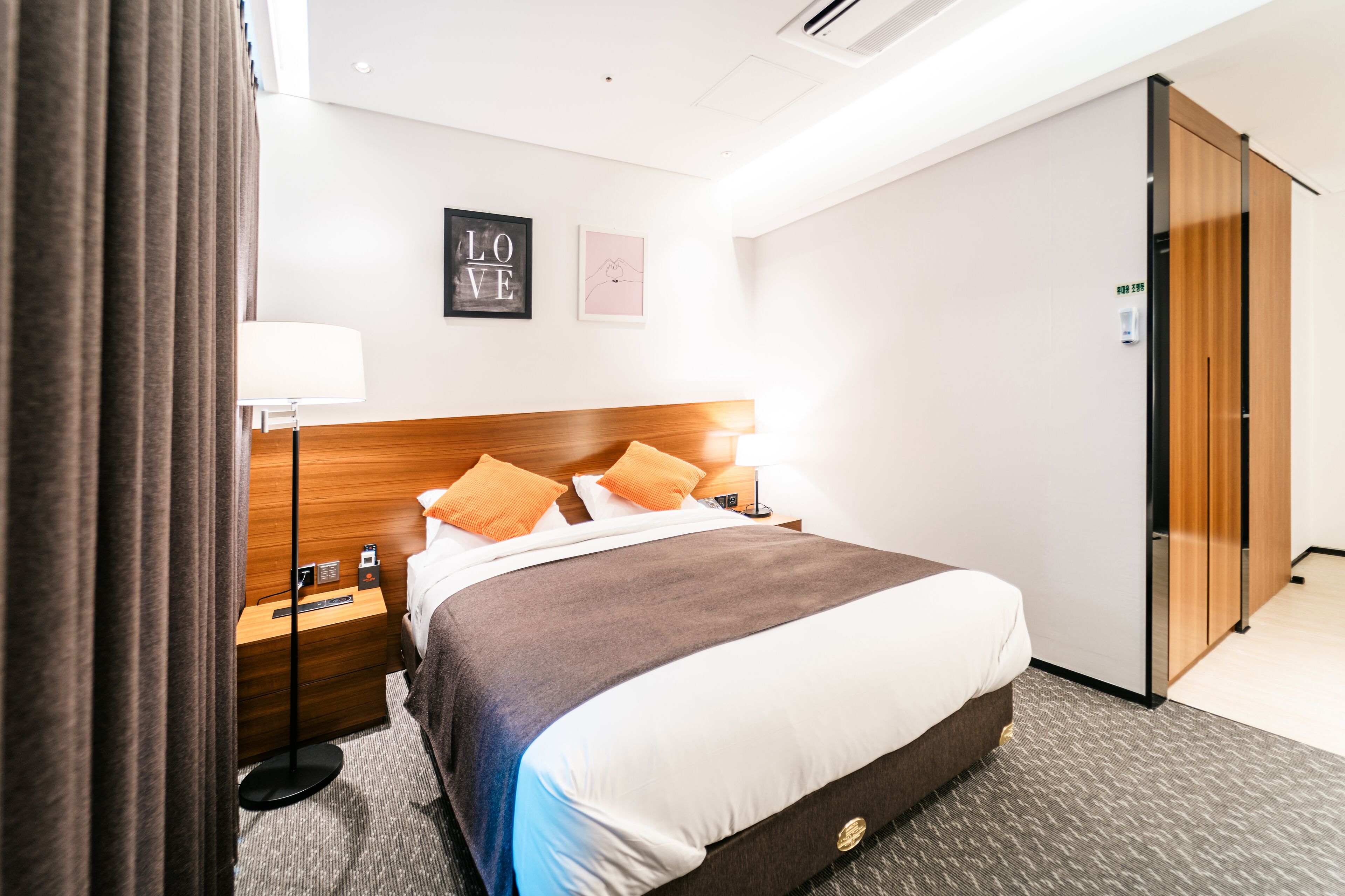 A hotel room with a wooden headboard, a bed with white and brown bedding, a nightstand with a lamp, and a wardrobe.