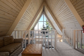 Une pièce mansardée chaleureuse, avec un plafond en pente en bois, une table à carreaux, un canapé à motifs et une vue sur l’extérieur par une fenêtre.