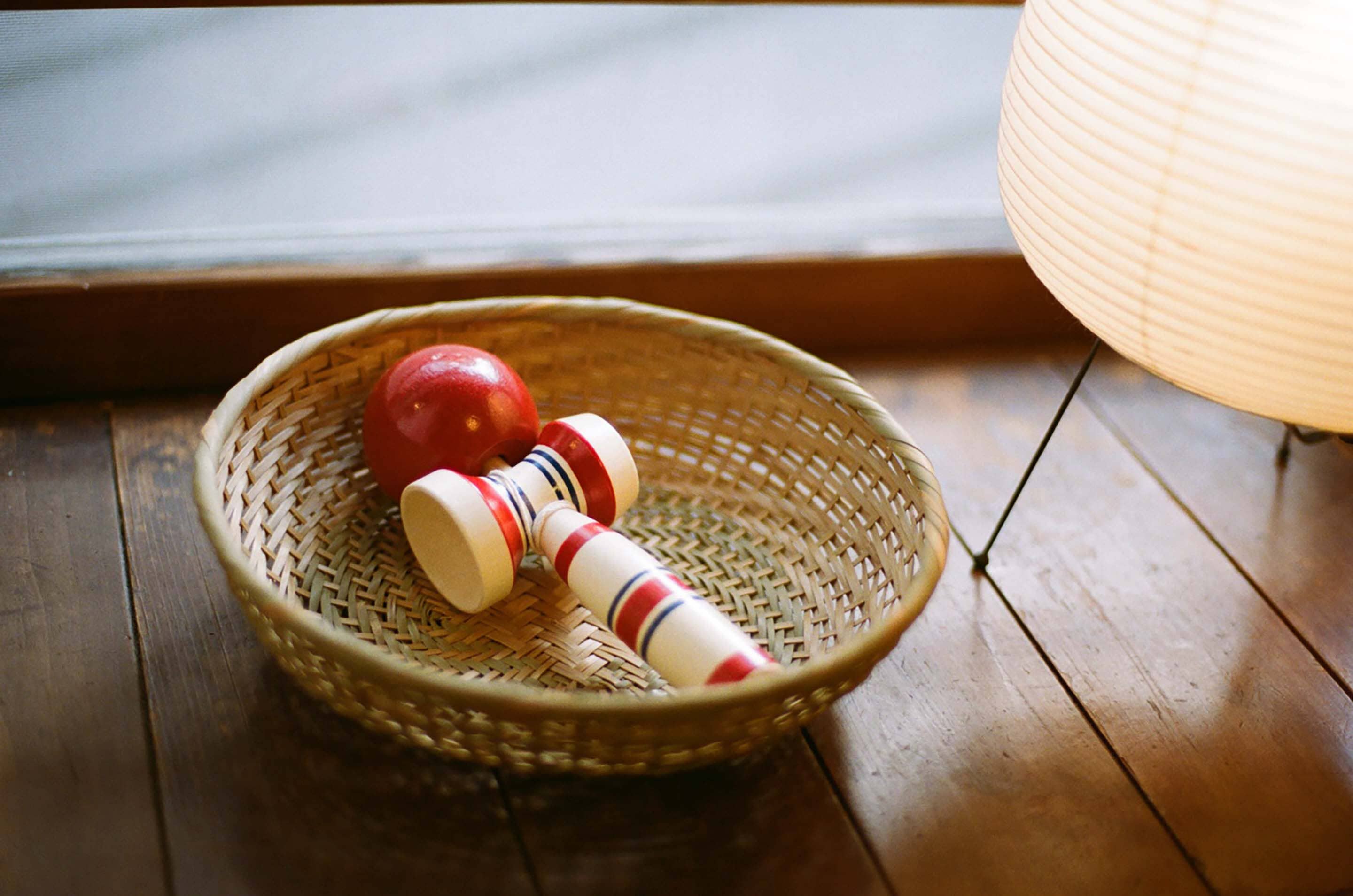 A woven basket containing a red apple and two wooden dumbbells on a wooden floor near a window with a light fixture.