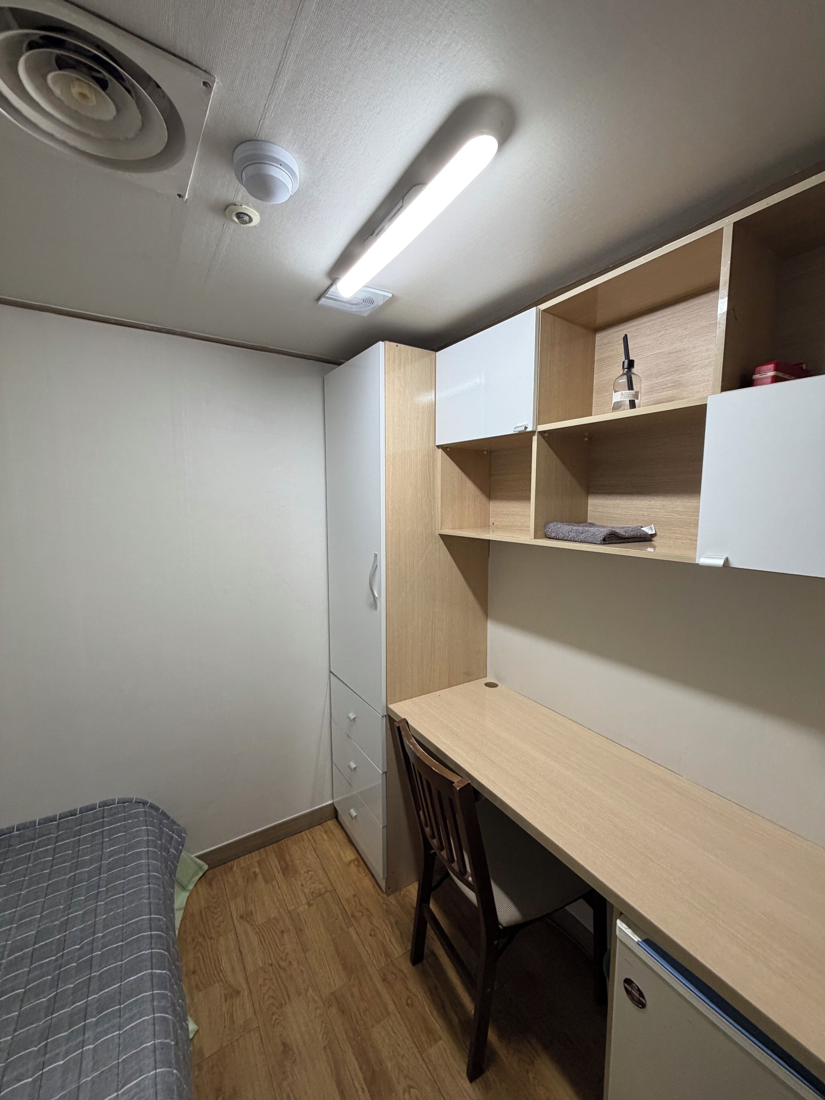 Une chambre à coucher de taille réduite, avec un lit simple, un bureau et une chaise. Des étagères intégrées se trouvent au-dessus du bureau et un petit placard en dessous.