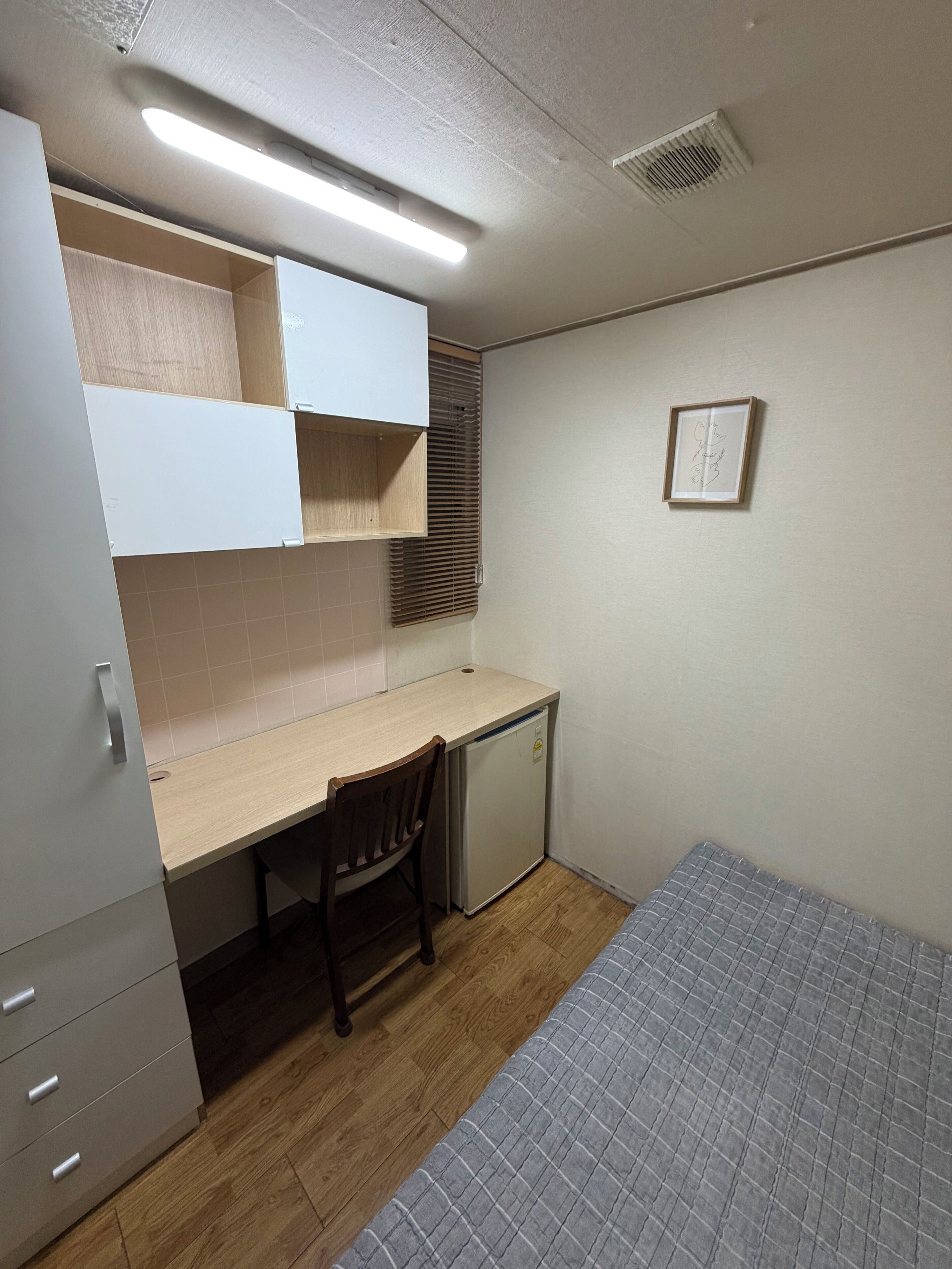 Una habitación compacta con una cama individual, un escritorio con silla, un refrigerador pequeño y un cuadro colgado en la pared.
