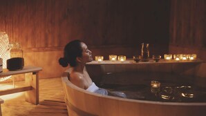 Uma pessoa relaxando em uma banheira de madeira, com velas e um pote de vidro sobre uma mesa de madeira.