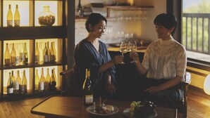 Deux personnes sont assises à une table en bois, devant des bouteilles de vin et des verres, dans une pièce où se trouve un meuble à vin et d’où l’on peut voir le paysage par une fenêtre.