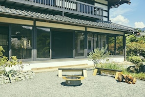 En traditionel bygning med store vinduer, en overdækket terrasse og et lille udendørs siddeområde med bænk og bålsted.