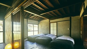 Une pièce de style japonais traditionnel, avec un sol recouvert de tatamis, un plafond bas avec des poutres en bois et une porte coulissante de type shoji. Trois tatamis sont disposés en rangée, accompagnés de coussins et d’un cadre de lit bas en bois.
