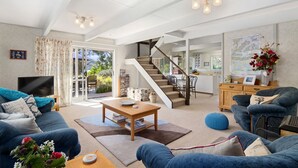 A spacious living room with a large sofa, a coffee table, and a TV. A staircase leads to a kitchen area with a dining table and chairs. - Pukenui Paradise - Havelock Holiday Home (Havelock)