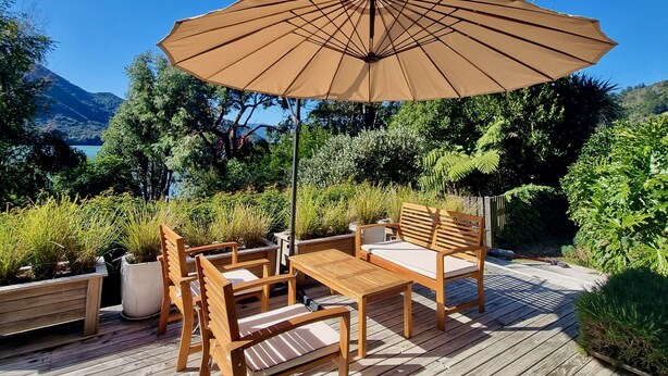 An outdoor wooden deck with a table and chairs, a large umbrella, and potted plants. - Pukenui Paradise - Havelock Holiday Home (Havelock)