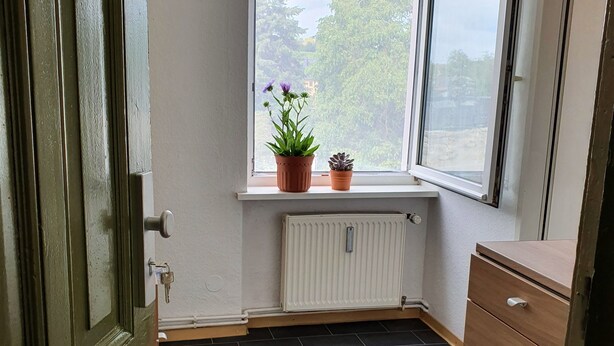 Una habitación en esquina con radiador, ventana con vistas a árboles, cómoda de madera y una puerta.