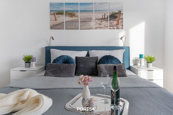 A neatly arranged bedroom with a bed, bedside tables, a tray with a bottle and glasses, and a beach-themed wall art.