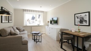 A modern living room with a sofa, a small table, a dining table with chairs, a TV mounted on the wall, and a window with a view of buildings.