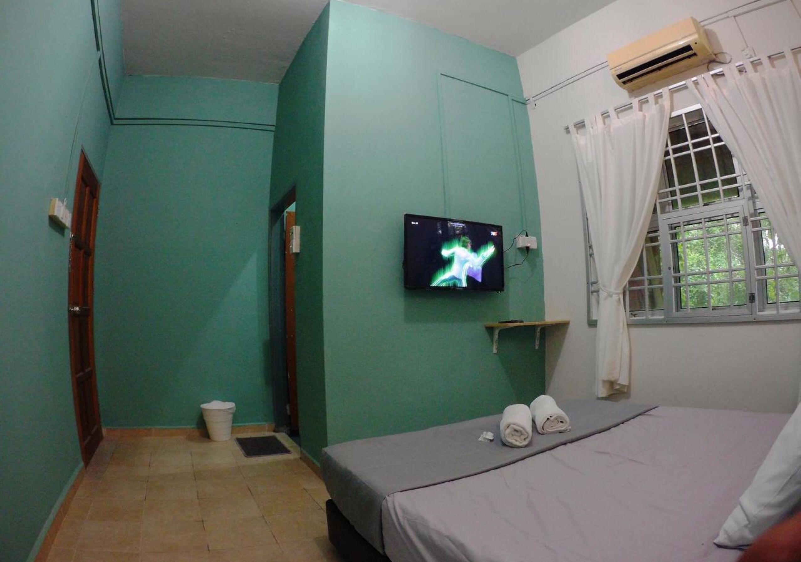 Een kamer met blauwgroene wanden, een bed met een grijze matras, een televisie aan de muur, een raam met witte gordijnen en een klein plankje.