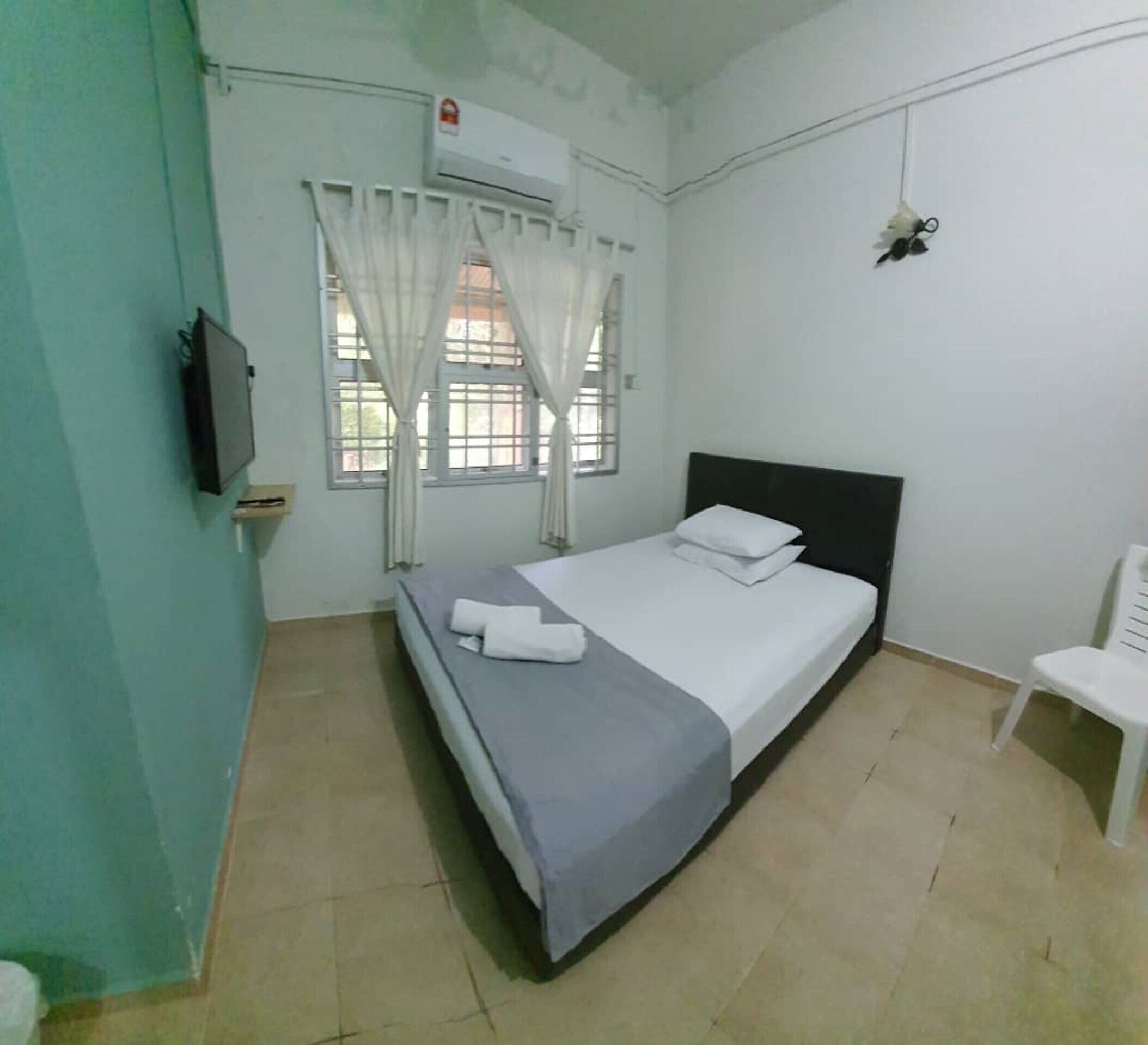A single-bed room with a grey bedspread, white pillows, a mounted TV, a window with white curtains, and a white chair.