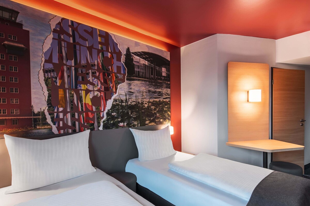 A hotel room with a large mural on the wall, a bed with white bedding, a bedside table with a lamp, and a wooden door.