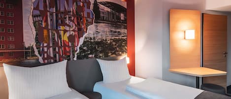 A hotel room with a large mural on the wall, a bed with white bedding, a bedside table with a lamp, and a wooden door.