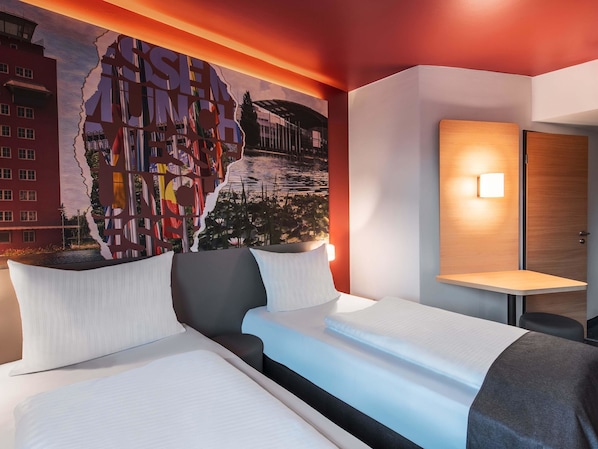 Una habitación de hotel con un gran mural en la pared, una cama con ropa blanca, una mesita de noche con una lámpara y una puerta de madera.