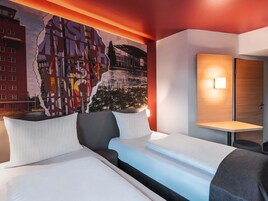 Una habitación de hotel con un gran mural en la pared, una cama con ropa blanca, una mesita de noche con una lámpara y una puerta de madera.