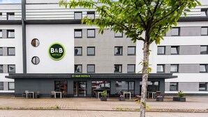 A B&B hotel with a modern facade, outdoor seating, and a tree in front. - B&B Hotel München-Messe (Aschheim)