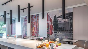 A modern dining area with a long white table, black bar stools, and a large window with a view of a building and greenery. - B&B Hotel München-Messe (Aschheim)