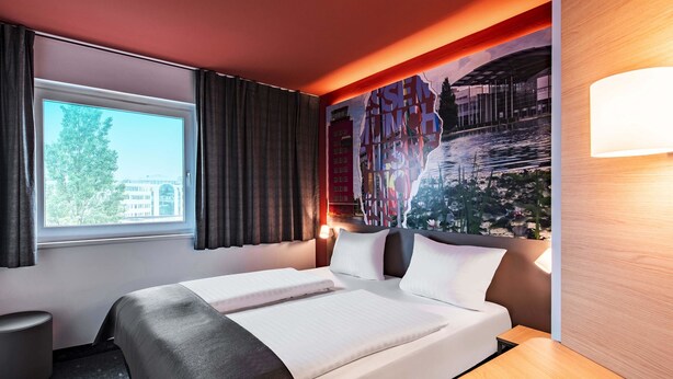 A hotel room with a bed, a window with curtains, a wall mural, and a bedside table. - B&B Hotel München-Messe (Aschheim)