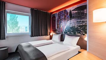 A hotel room with a bed, a window with curtains, a wall mural, and a bedside table.