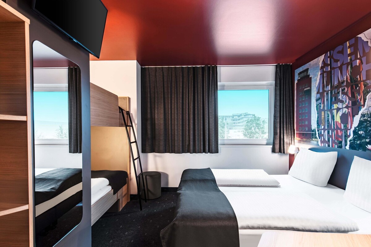 A modern hotel room with a bunk bed, a desk, and a large window offering a city view.
