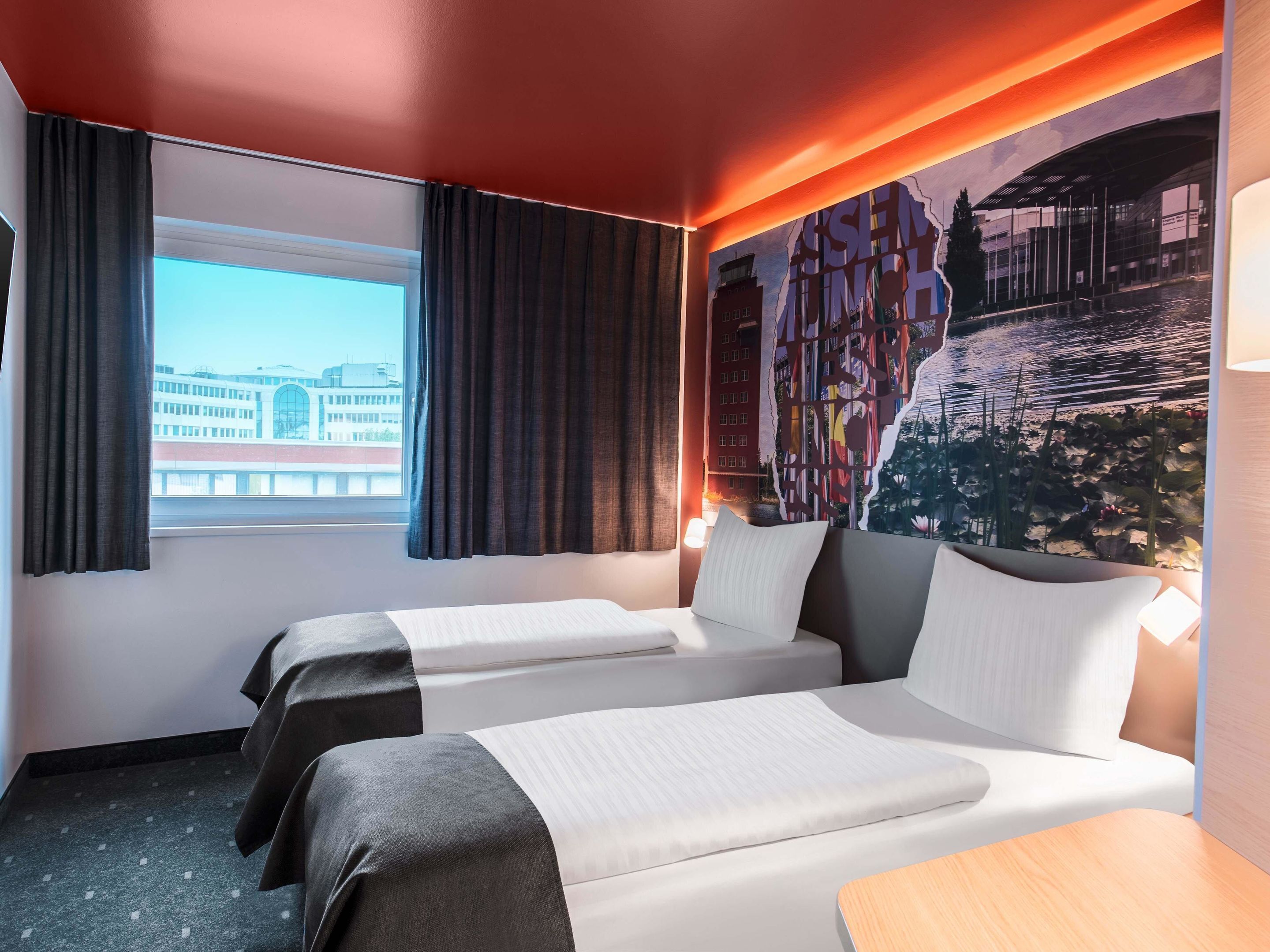 A hotel room with two beds, a large window with curtains, a cityscape mural on the wall, and a lamp on the nightstand.