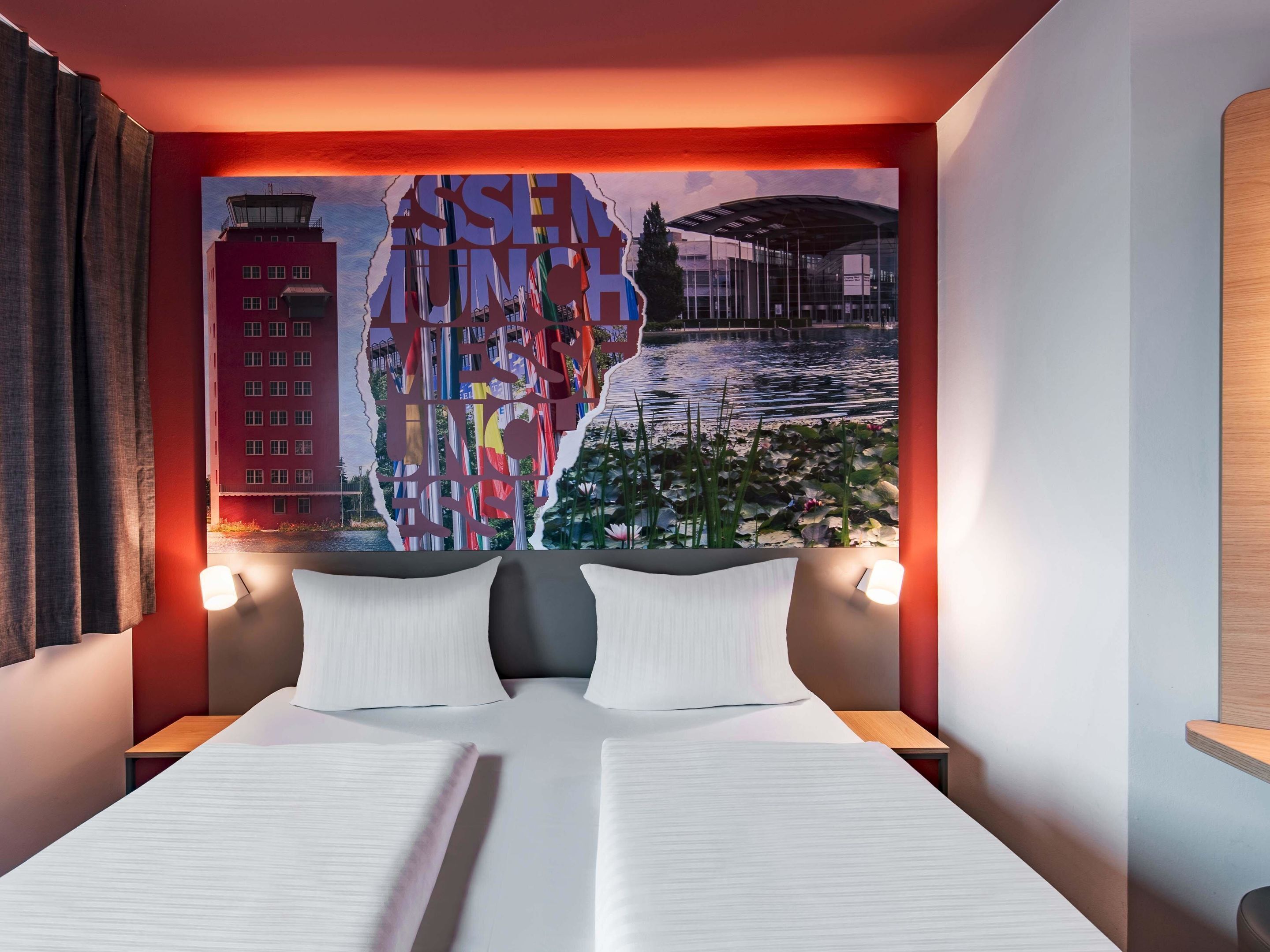 A hotel room with a bed, two bedside tables, a wall mural, and a view of a building and water.