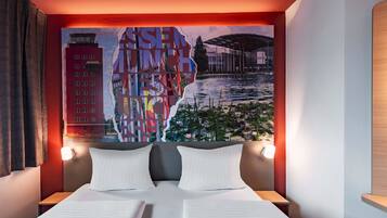 A hotel room with a bed, two bedside tables, a wall mural, and a view of a building and water.
