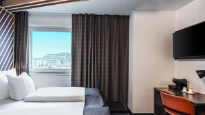 A hotel room with a bed, a desk, a chair, a TV, and a window with curtains. - B&B Hotel München-Messe (Aschheim)