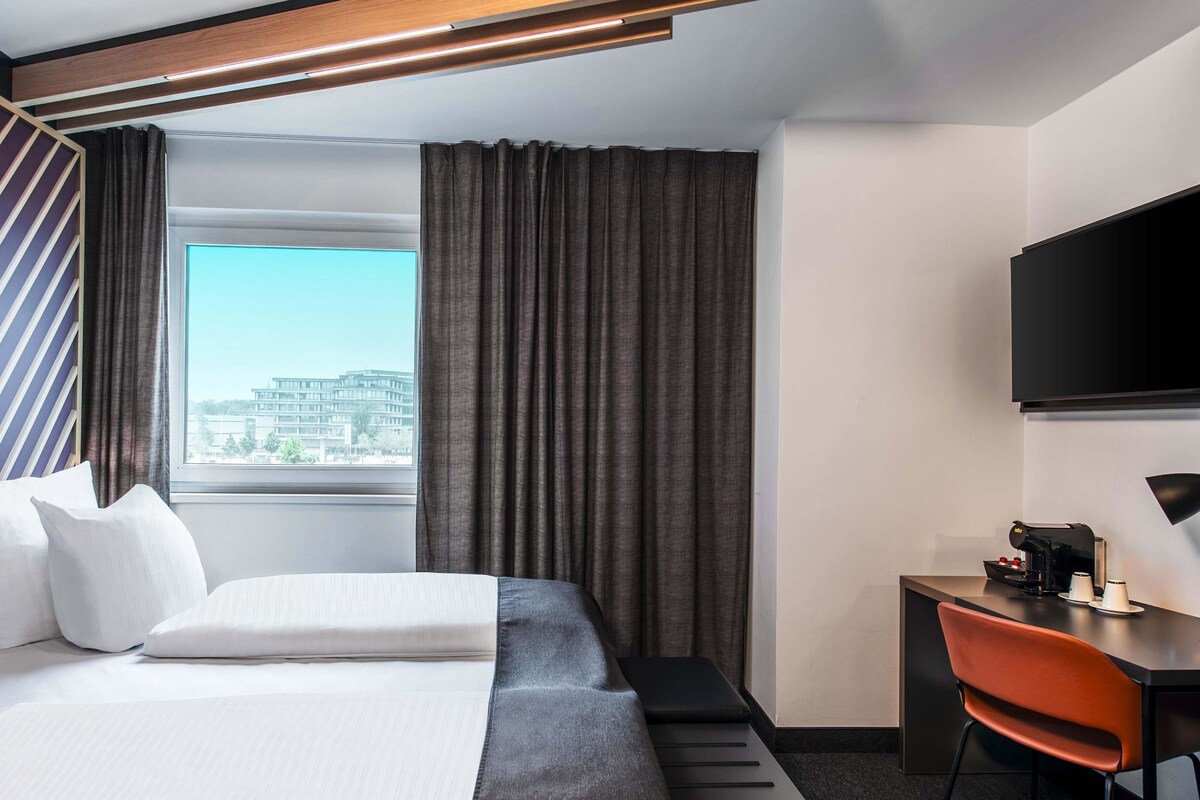 A hotel room with a bed, a desk, a chair, a TV, and a window with curtains.