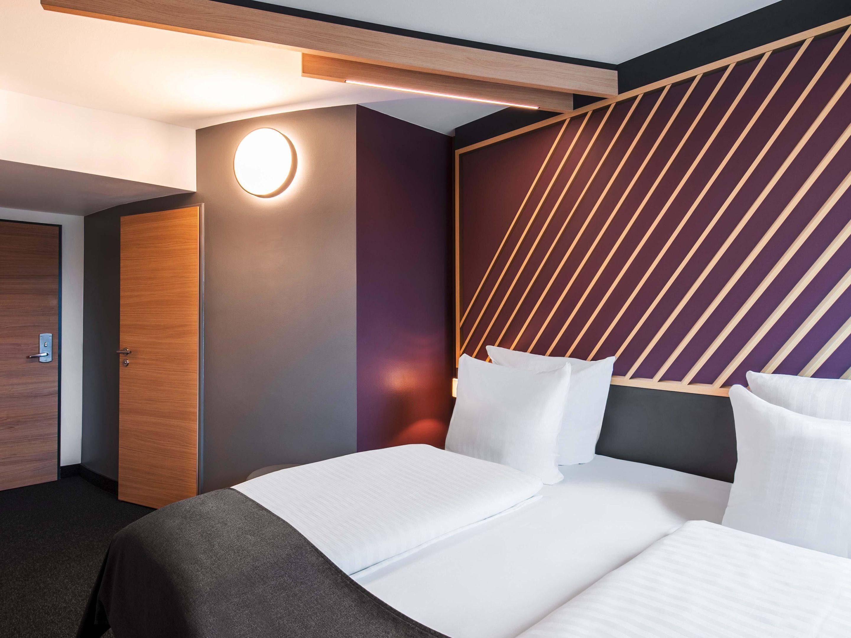 A hotel room with a modern design, featuring a large bed with white linens, a wooden headboard with diagonal lines, and a circular light fixture on the wall.