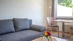 A modern living room with a grey sofa, a wooden coffee table with a vase of flowers, a dining table with chairs, and a window with curtains. - Ferienhaus Anja - FH Anja (Plau am See)