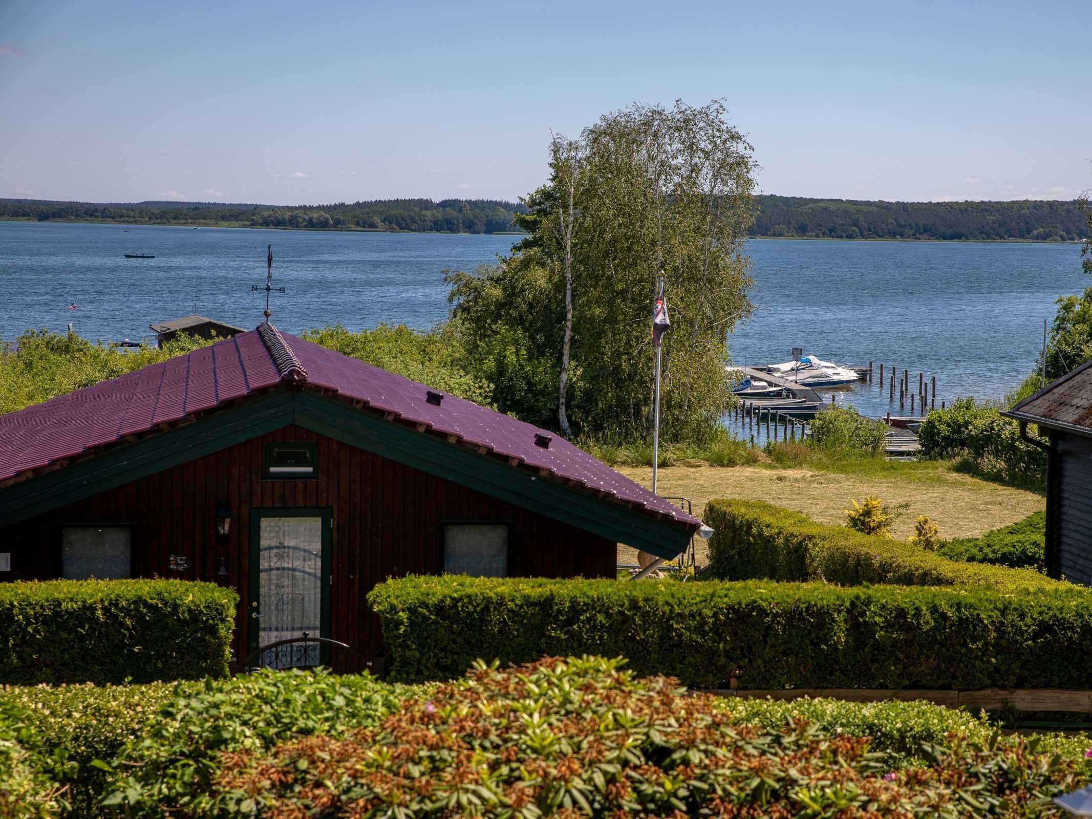 A rustic cabin with a red roof, surrounded by well-maintained hedges and a view of a calm lake with boats docked at a pier.