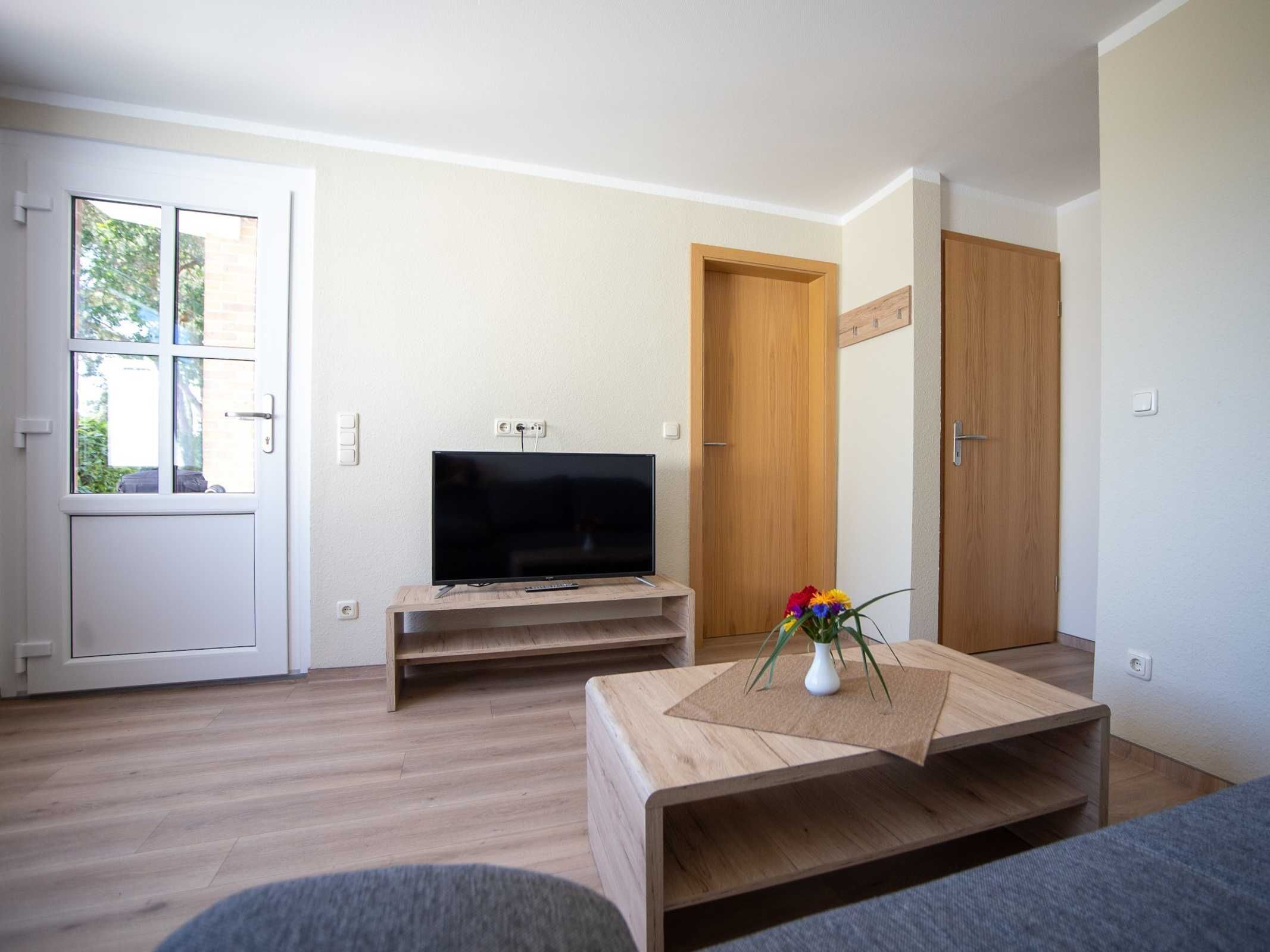 A modern living room with a flat-screen TV on a wooden stand, a small coffee table with a vase of flowers, and a door leading to another room.